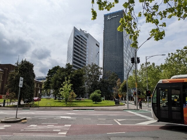 A photo of some of the Melbourne skyline from street level with the front of a bus in the foreground that seems to have no wheels